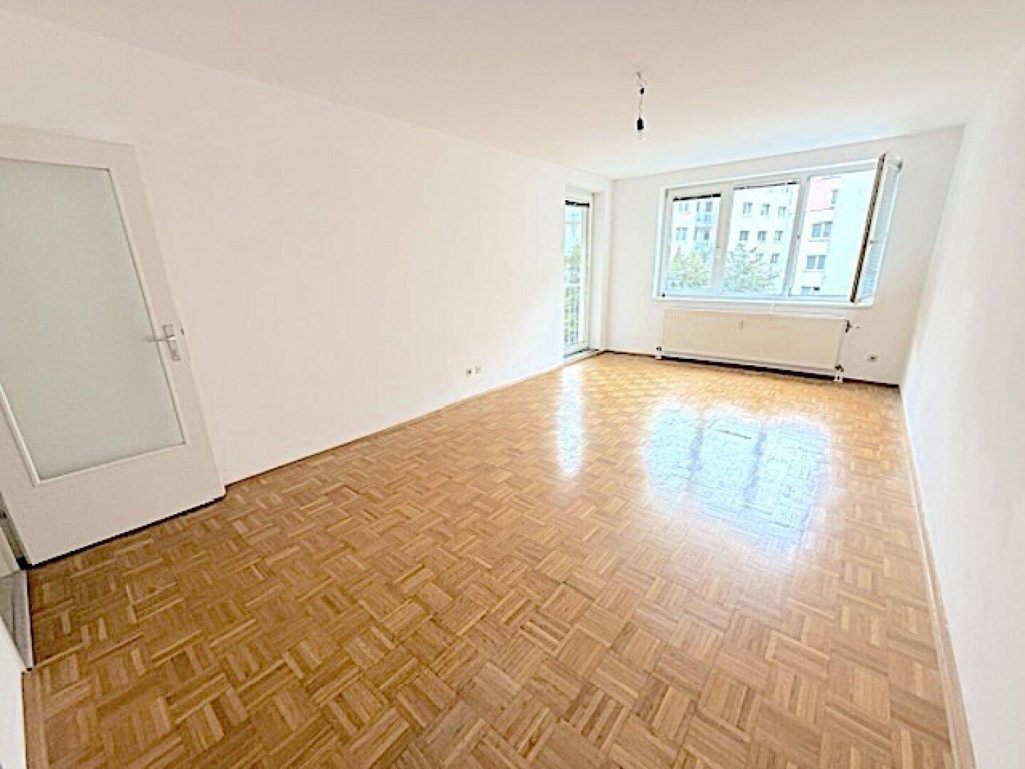 Geräumiges, leeres Wohnzimmer mit Fischgrätparkett und Zugang zum Balkon.