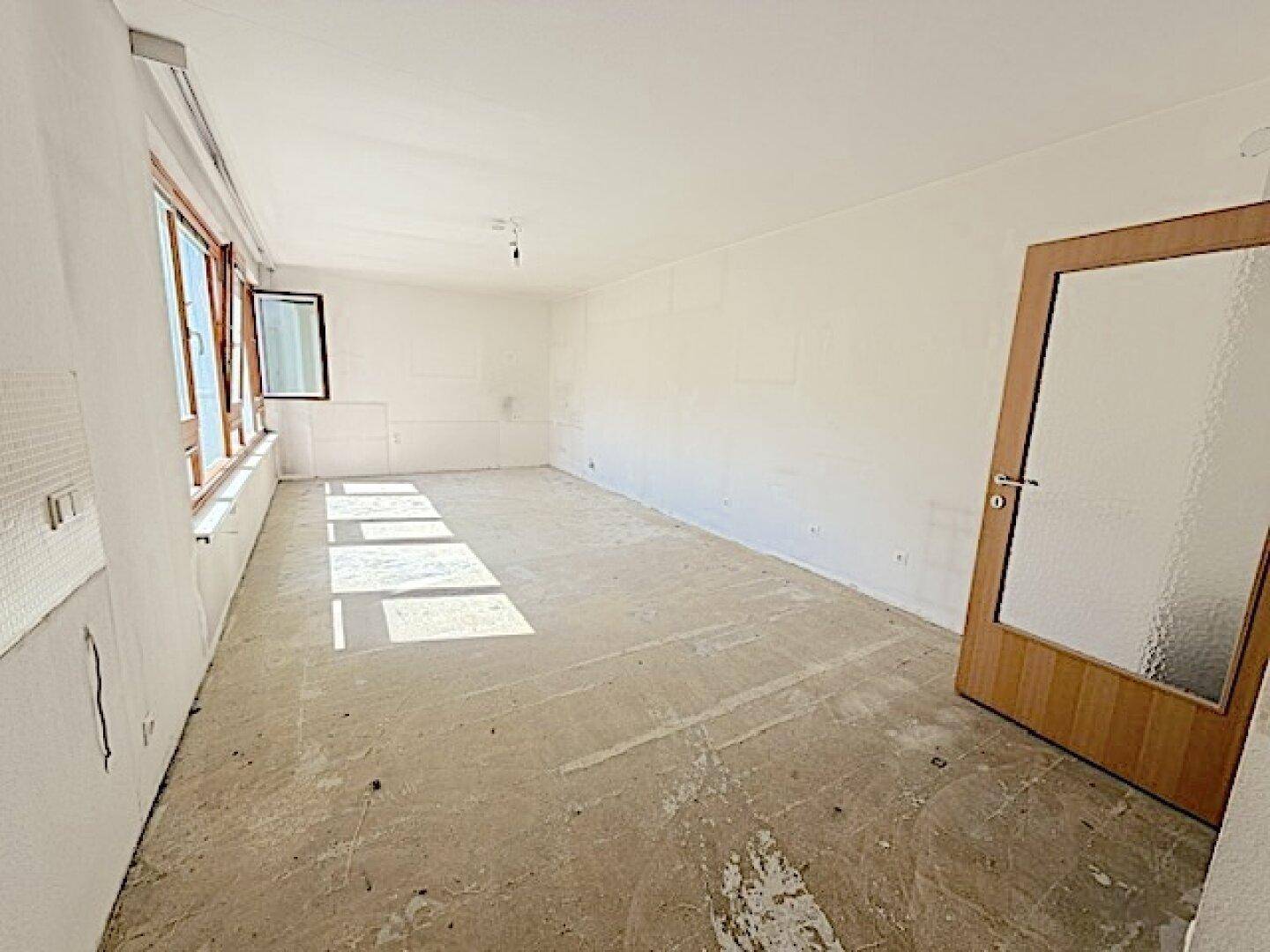 Unmöblierter Wohnbereich mit Betonboden, großen Fenstern und einer Holztür, bereit für die Fertigstellung.