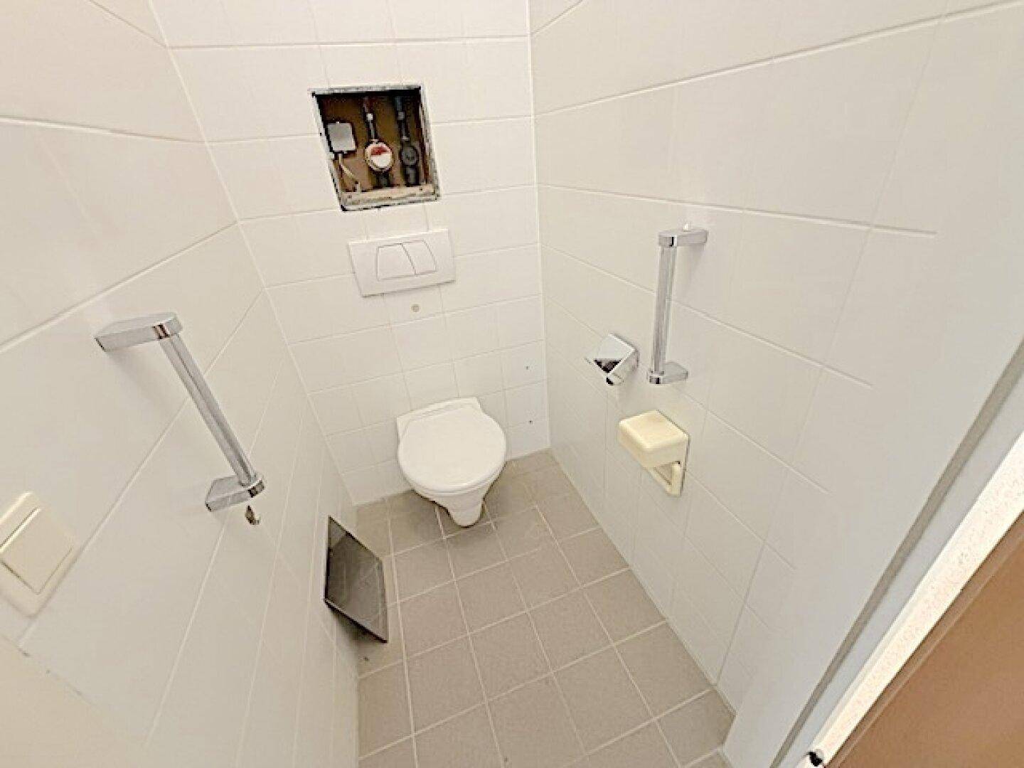 Separates WC mit Wand-WC, weißen Fliesen und praktischen Haltegriffen für Barrierefreiheit.