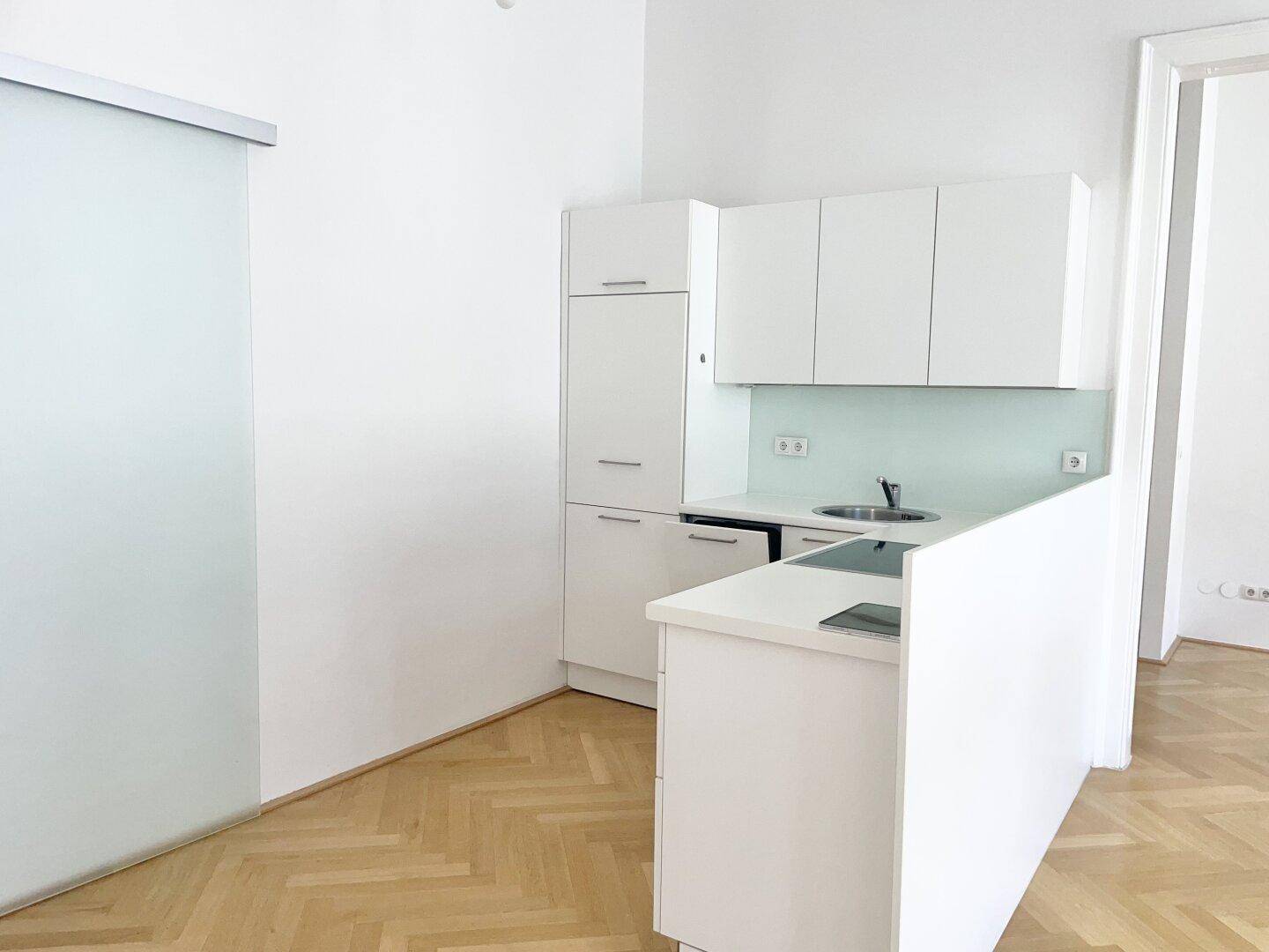 Moderne Küchenzeile mit weißen Schränken, Spüle und Induktionskochfeld, daneben eine Glasschiebetür.