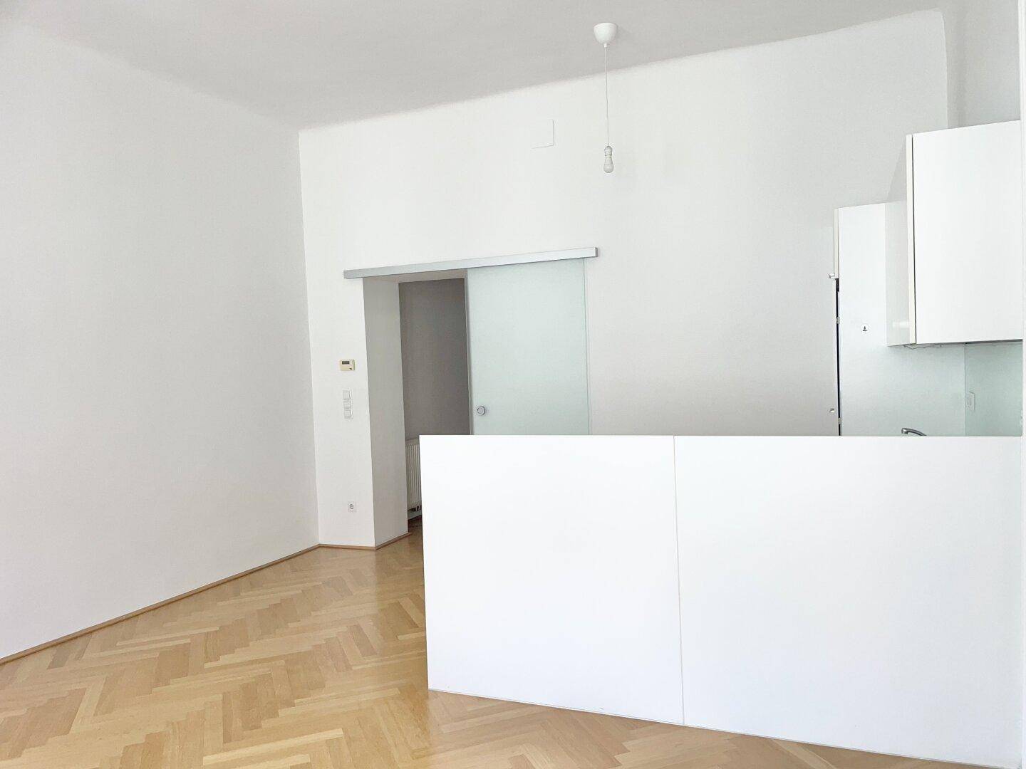 Offener Wohnbereich mit Fischgrätparkett und einer modernen Glasschiebetür zu einem angrenzenden Raum.