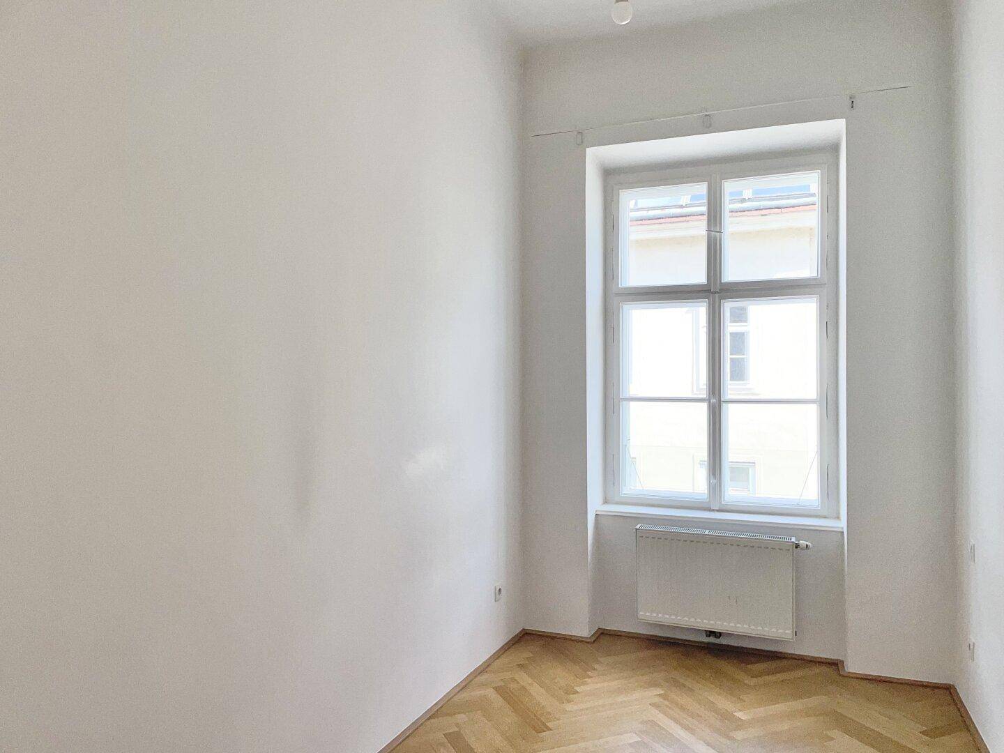 Helles Schlafzimmer mit Fischgrätparkett und einem großen Fenster, ideal für Ruhe und Entspannung.