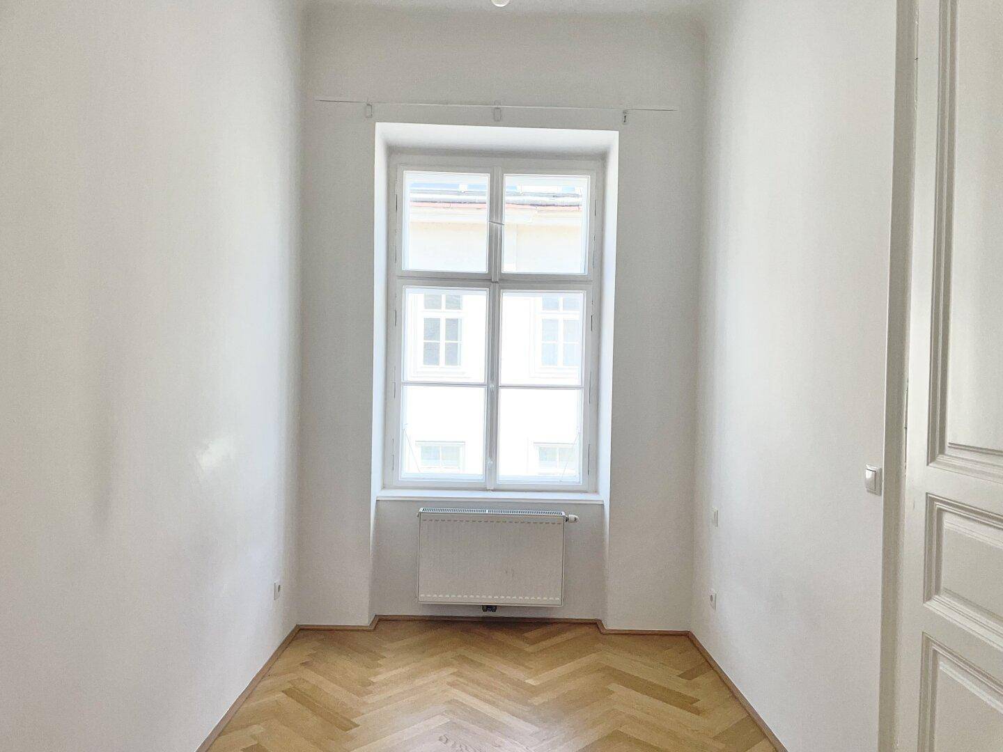 Gemütliches Schlafzimmer mit Fischgrätparkett, einem großen Fenster und einer klassischen Holztür.