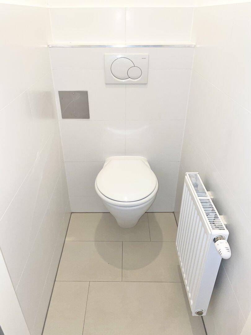 Modernes WC mit wandhängender Toilette, weißen Fliesen und einem Heizkörper für Komfort.
