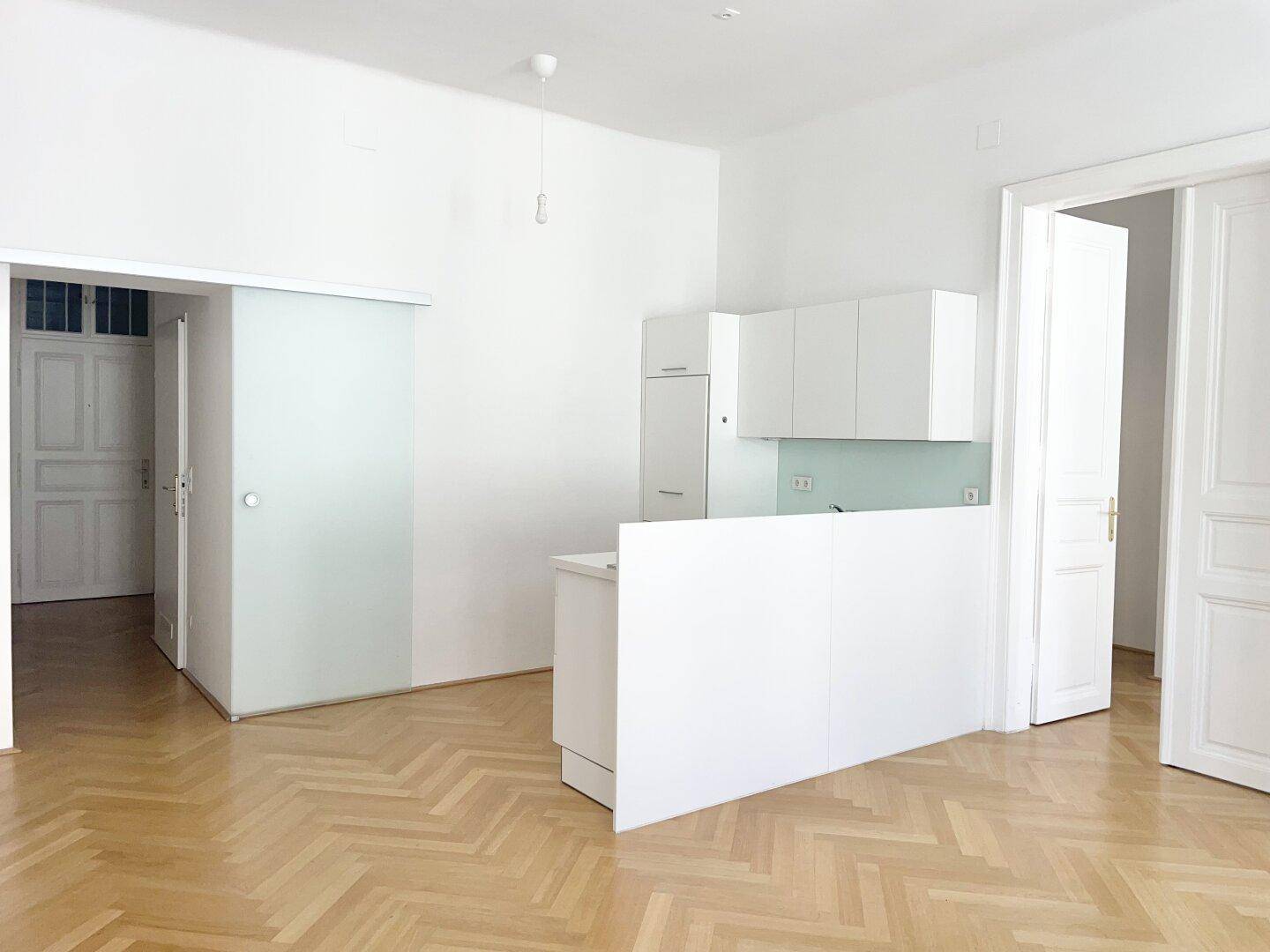 Geräumiger Wohnbereich mit integrierter Küchenzeile, Fischgrätparkett und eleganten Doppeltüren.