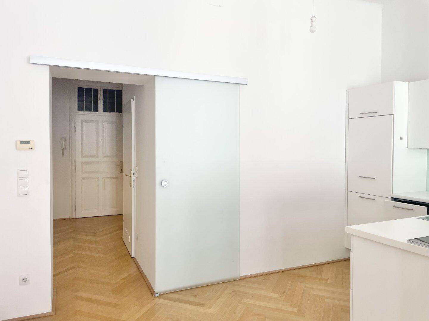 Moderne Glasschiebetür, die zu einem Flur mit traditioneller Eingangstür und Gegensprechanlage führt.