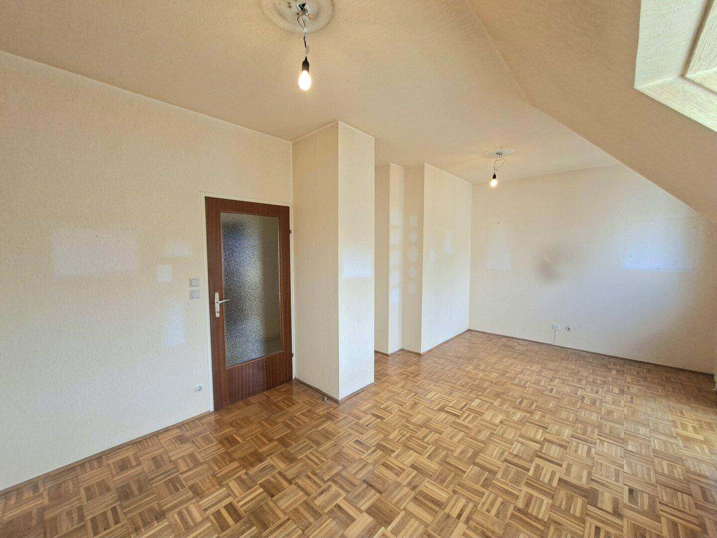 Geräumiger Raum mit Parkettboden, Dachschräge und einer Holztür.