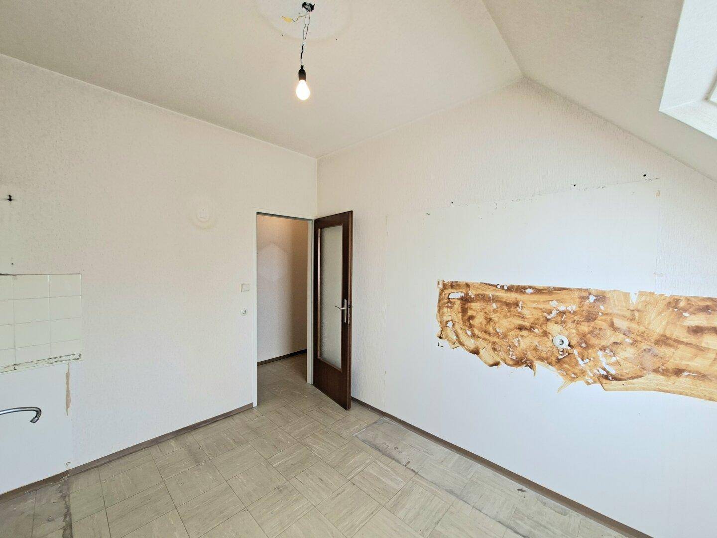 Leere Küche mit alten Wandfliesen, Holztür und sichtbaren Renovierungsspuren.