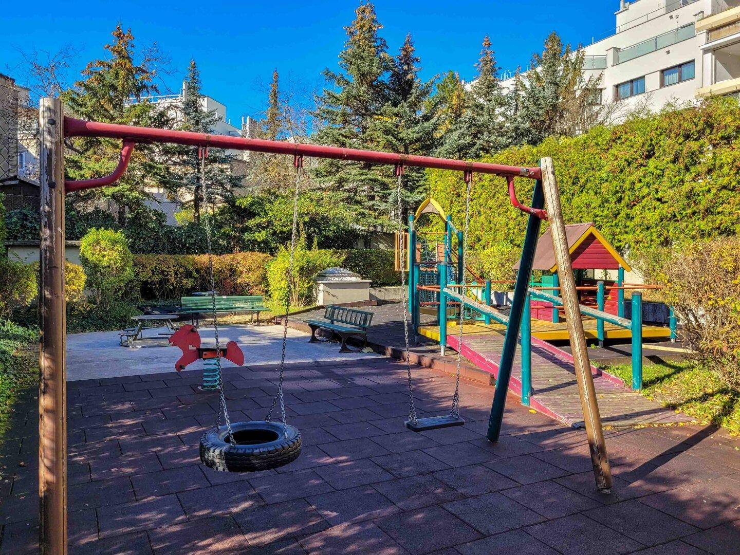 Bunter Kinderspielplatz mit Schaukeln, Rutsche und weichem Bodenbelag, umgeben von Bäumen.