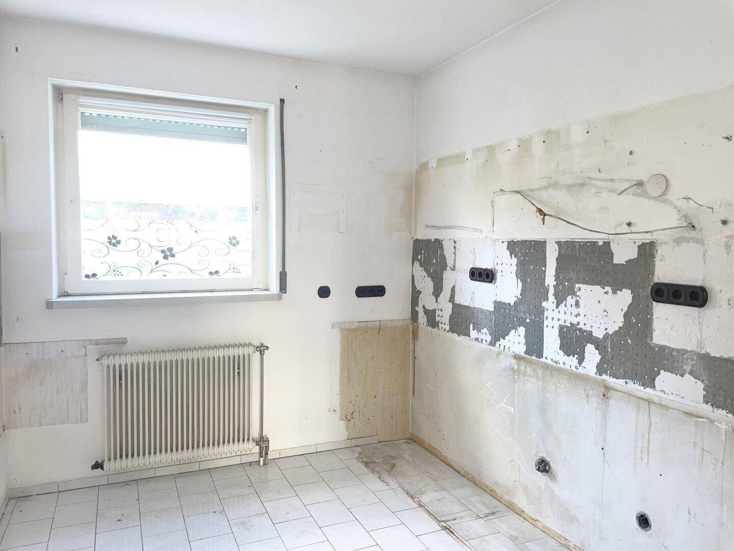 Leere Küche mit Fliesenboden, Fenster und Heizkörper, bereit für eine neue Einbauküche.