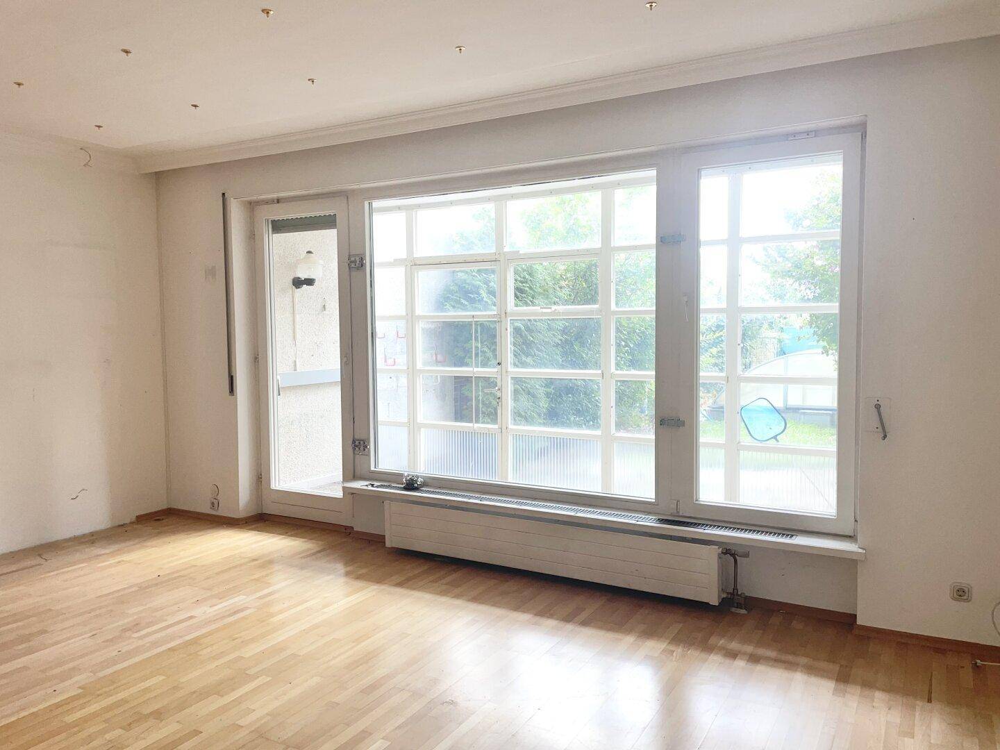Helles, leeres Wohnzimmer mit Parkettboden, großen Fenstern und Heizkörpern.