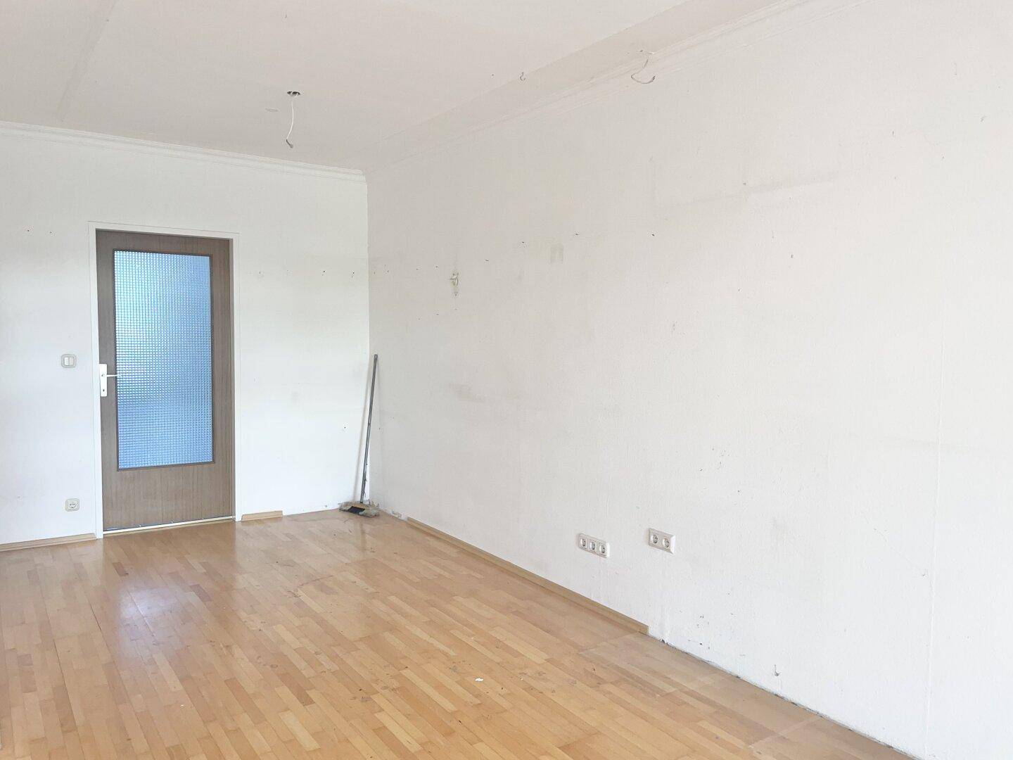 Leeres Wohnzimmer mit hellem Parkettboden und einer Tür, die zu weiteren Räumen führt.