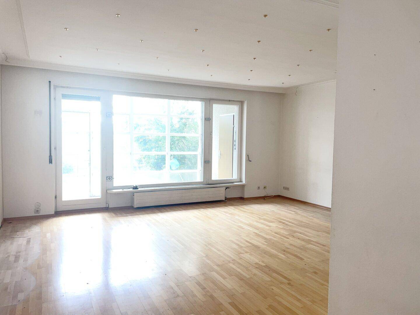 Lichtdurchflutetes Wohnzimmer mit Parkettboden und großen Fenstern, die viel Tageslicht hereinlassen.