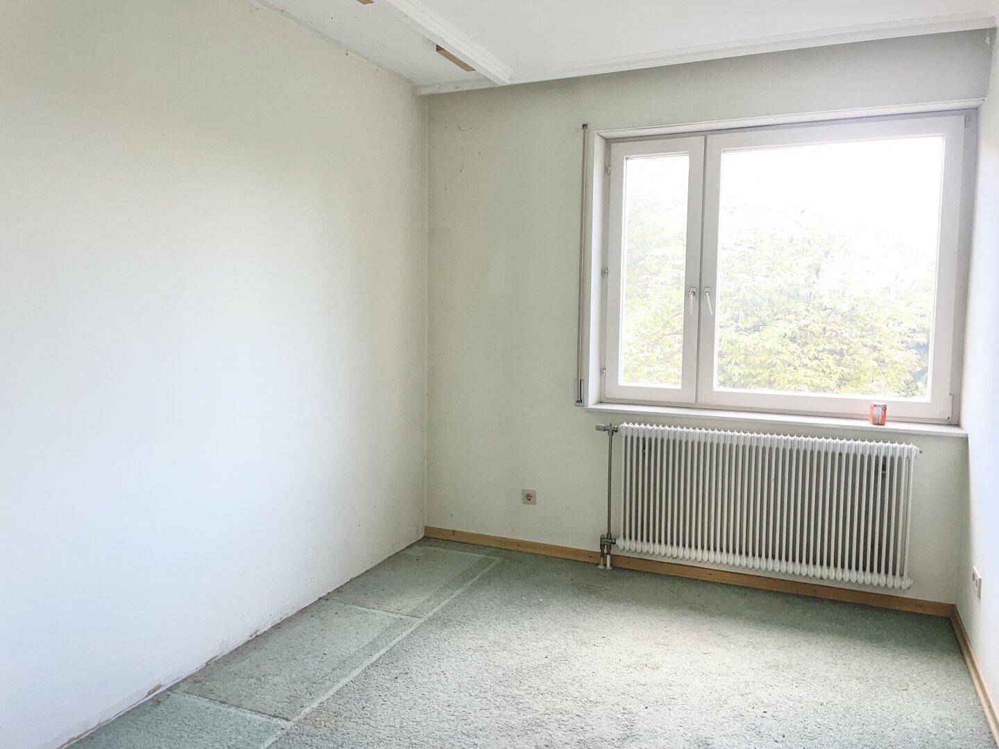 Leeres Schlafzimmer mit Fenster, Heizkörper und grünem Teppichboden.