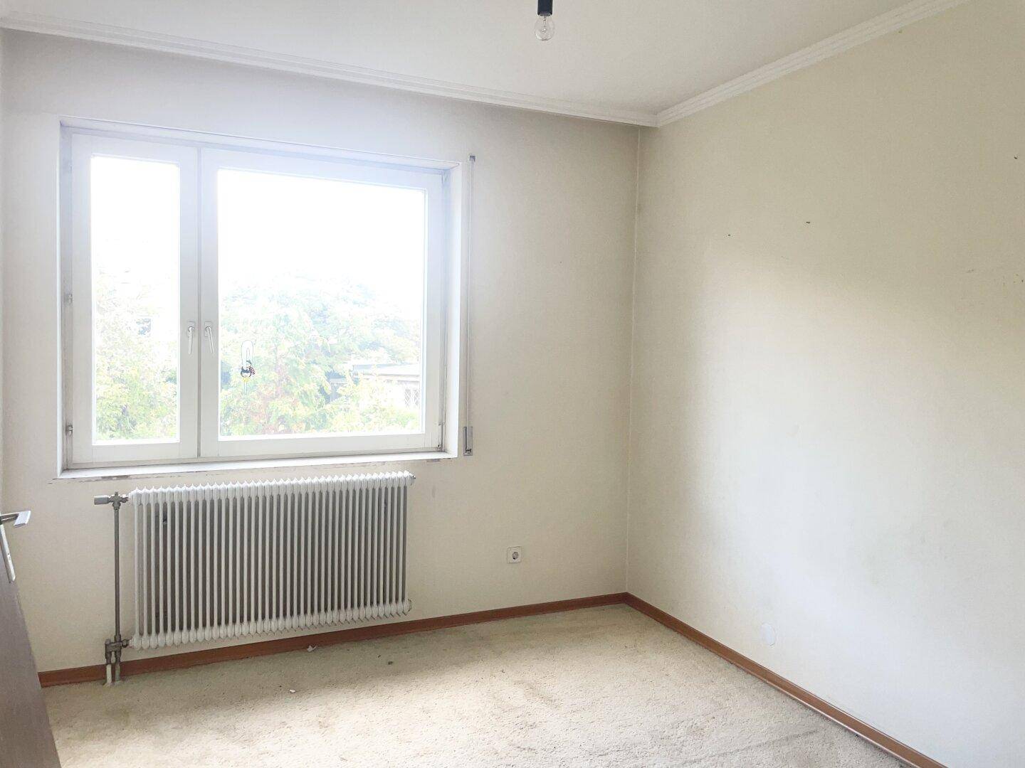 Helles Schlafzimmer mit Fenster, Heizkörper und hellem Teppichboden.