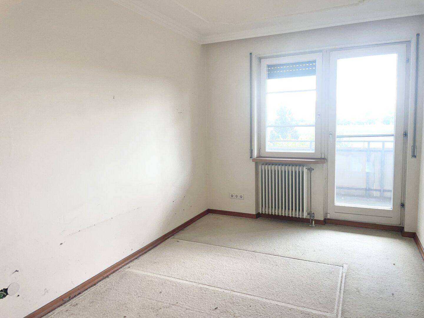 Schlafzimmer mit Fenster, Heizkörper und Teppichboden, mit Zugang zu einem Balkon.