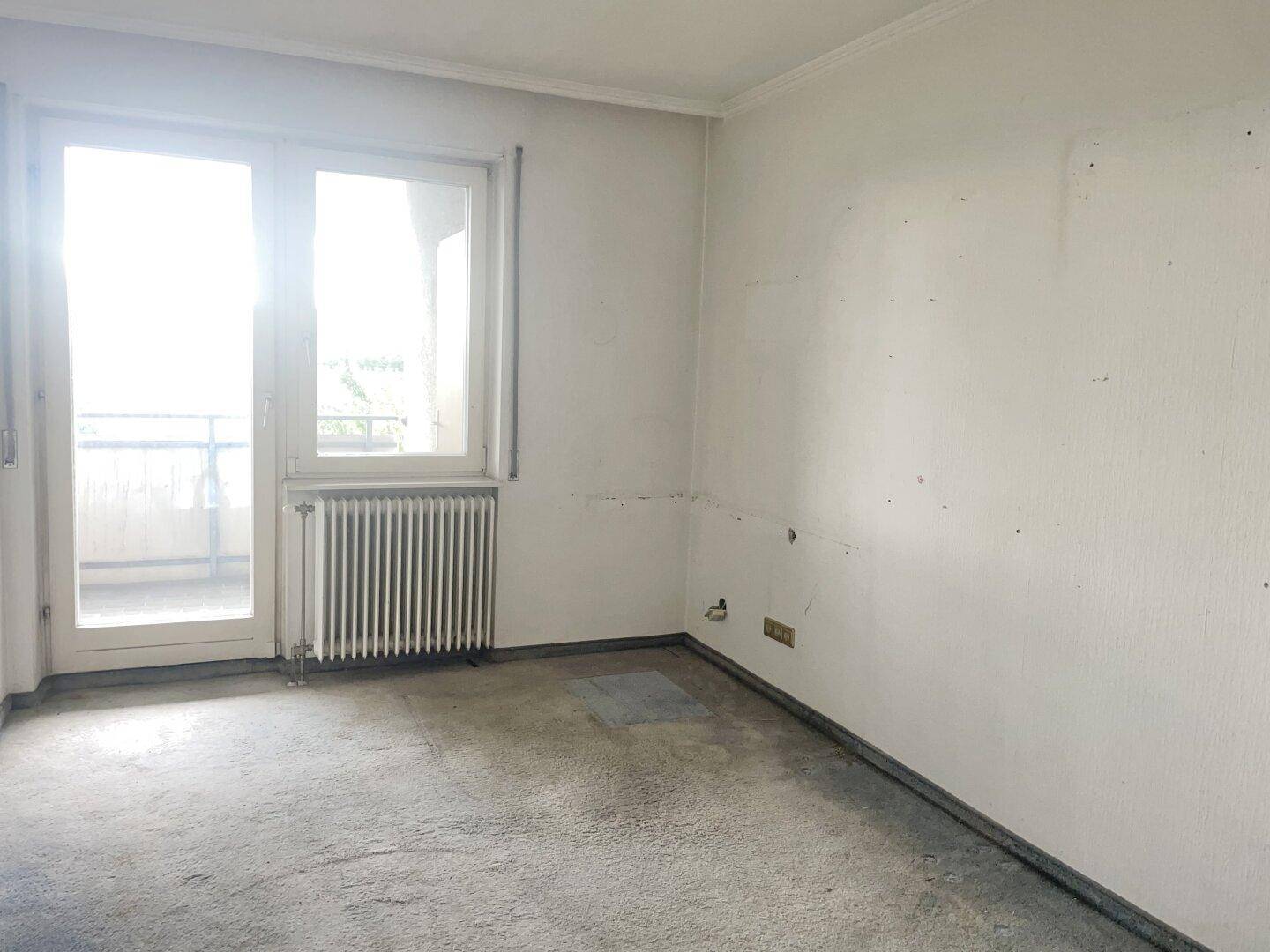 Schlafzimmer mit Fenster, Heizkörper und grauem Teppichboden, mit Zugang zu einem Balkon.