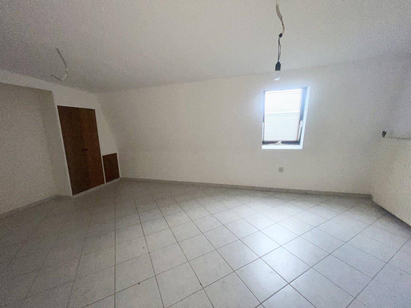 Leeres Dachzimmer mit Dachfenster, Fliesenboden und schräger Decke.