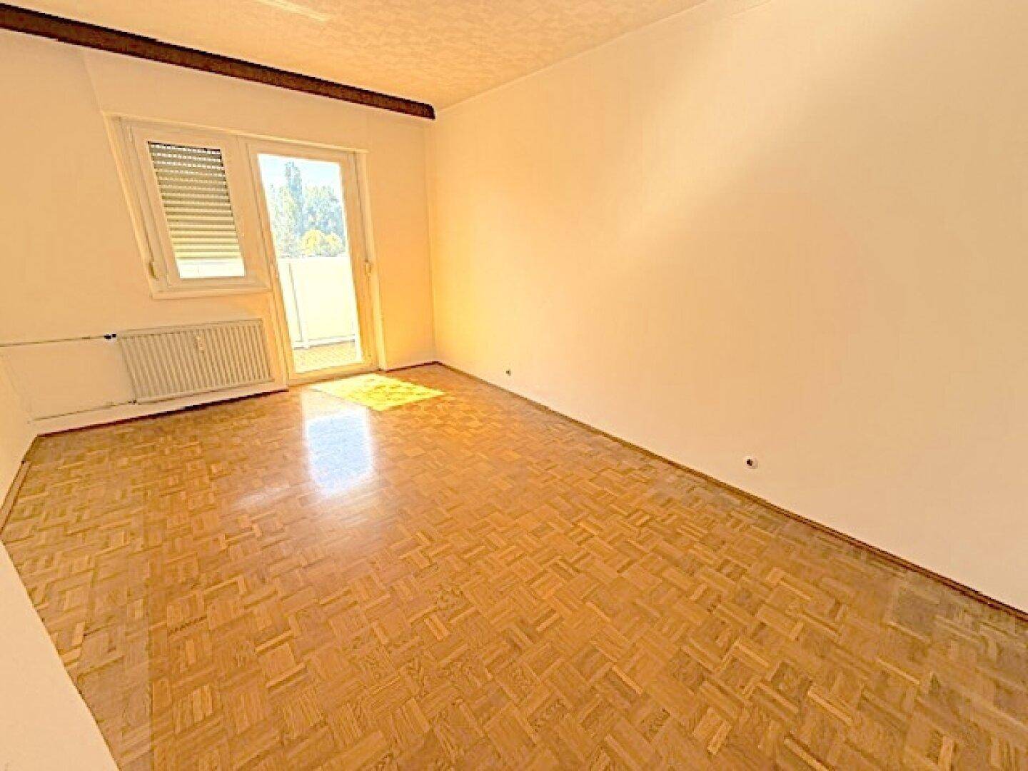 Leerer Raum mit Parkettboden und Zugang zu einem sonnigen Balkon, der viel Licht spendet.