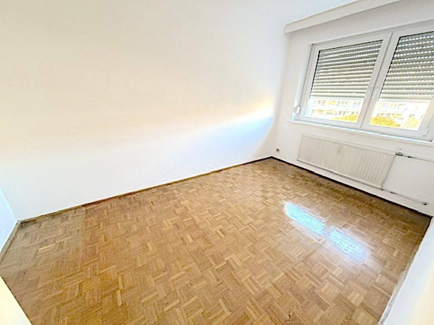 Leeres Zimmer mit Parkettboden und Fenster mit Rollläden, das viel Licht hereinlässt und Privatsphäre bietet.