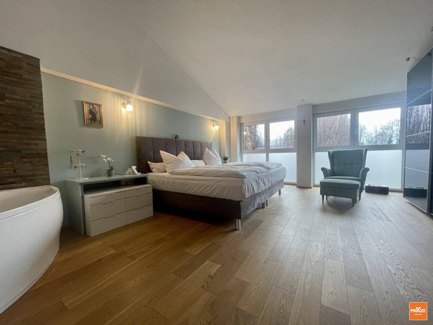 Großes Schlafzimmer mit Holzboden, breiten Fenstern und integrierter Badewanne.