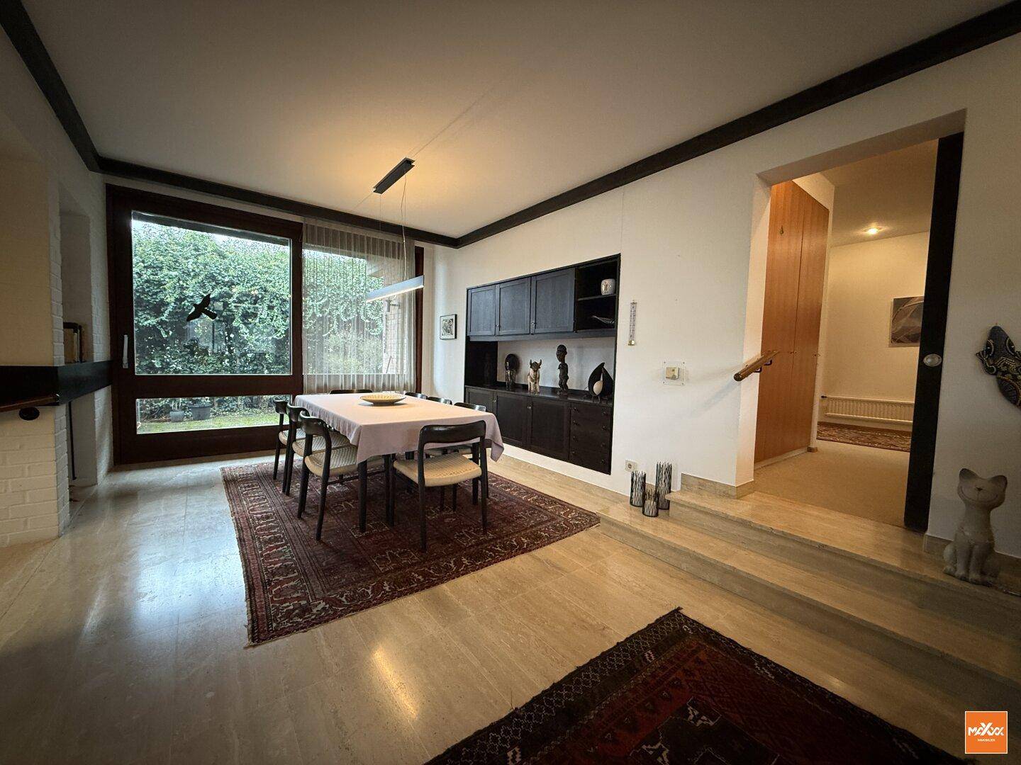 Esszimmer mit großem Fenster zum Garten, dunklen Einbauschränken und Teppich auf hellem Fliesenboden.