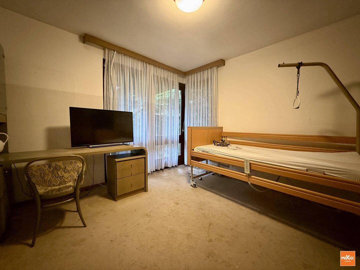 Schlafzimmer mit Pflegebett, Schreibtisch und Fenster mit Vorhängen, das Tageslicht hereinlässt.