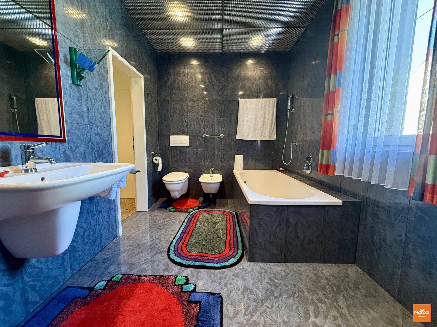 Geräumiges Badezimmer mit dunklen Fliesen, Badewanne, Bidet und Fenster.
