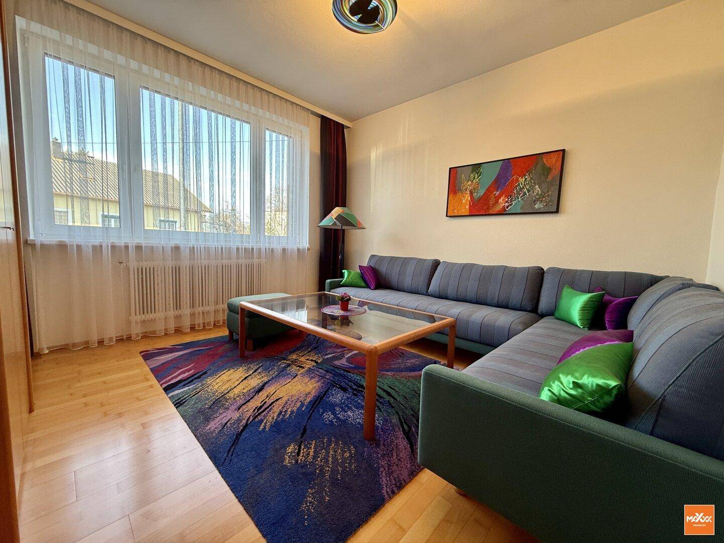 Helles Wohnzimmer mit großem Fenster, Teppich und bequemer Sofagarnitur.