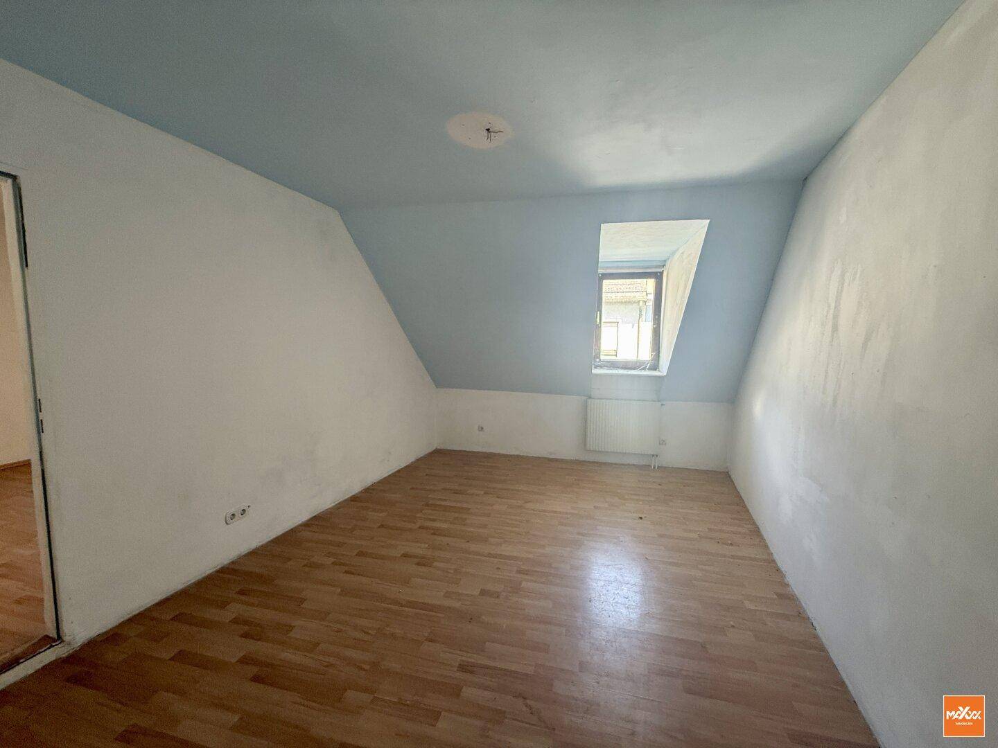Helles Schlafzimmer mit Dachschräge, Holzboden und einem Fenster, das viel Tageslicht hereinlässt.