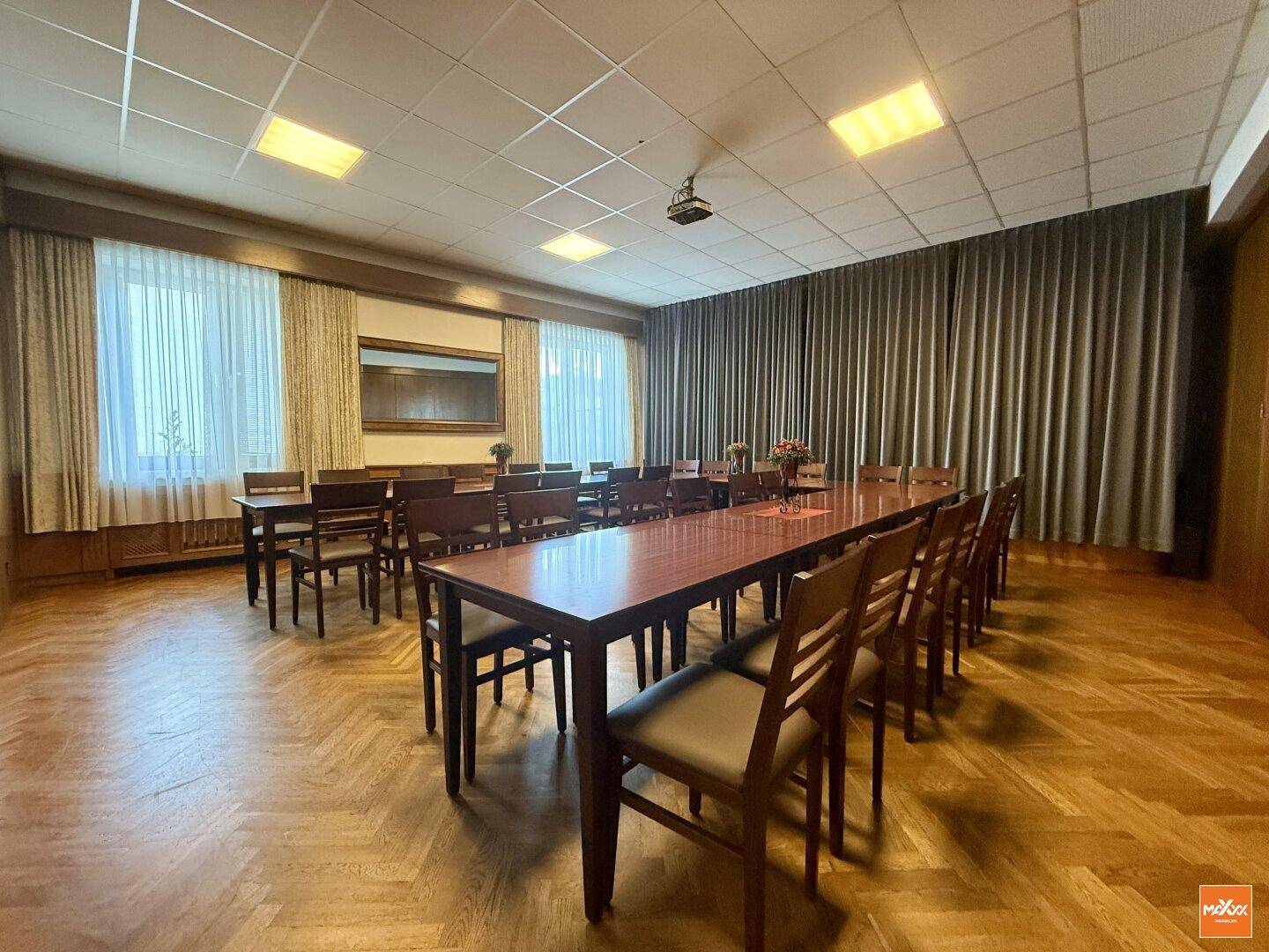 Konferenz- oder Speisesaal mit Holztischen, Vorhängen und Projektor an der Decke.