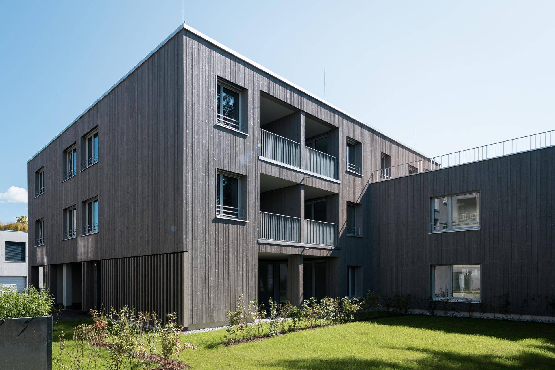 Ansicht eines modernen Wohngebäudes mit Holzverkleidung und Balkonen, umgeben von Grünflächen und Rasen.