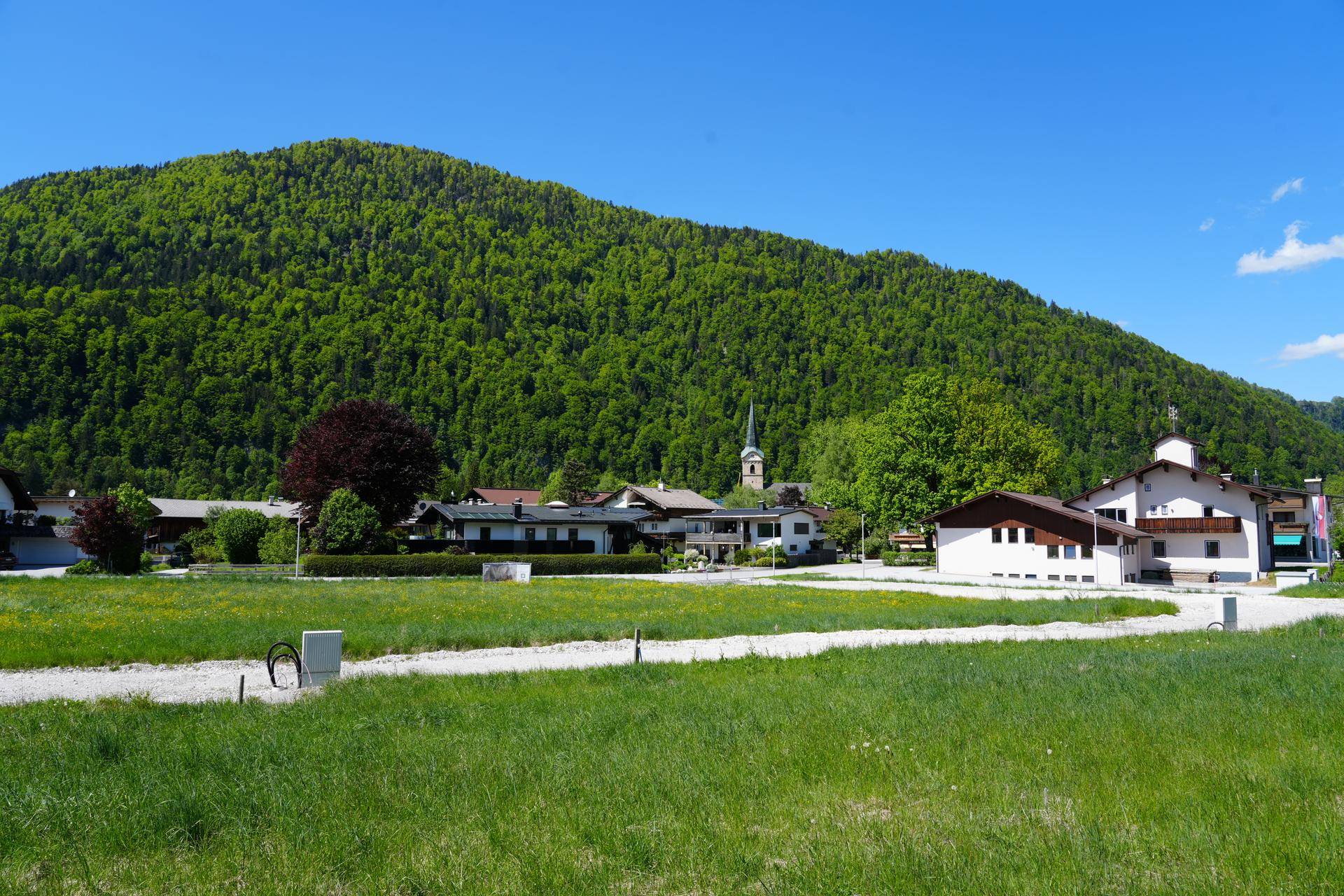 Idyllische Dorfansicht mit traditionellen Häusern, grünen Wiesen und einem bewaldeten Berg im Hintergrund.