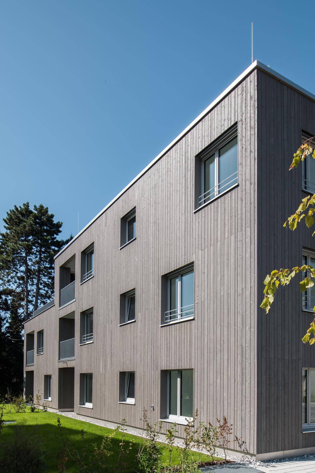 Seitenansicht des modernen Wohnhauses mit vertikaler Holzfassade unter blauem Himmel und Bäumen.