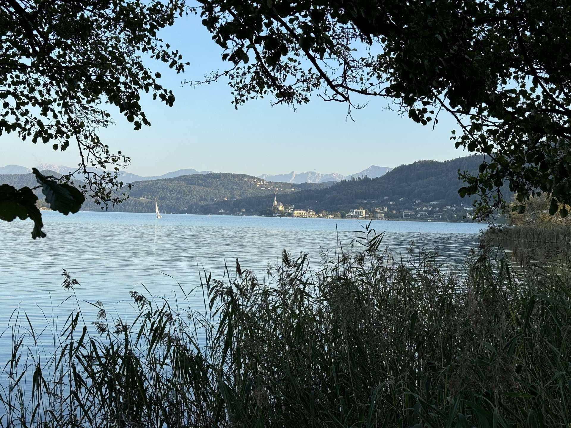 Ruhiger See mit Schilf im Vordergrund und Bergen sowie einem Dorf am Ufer.