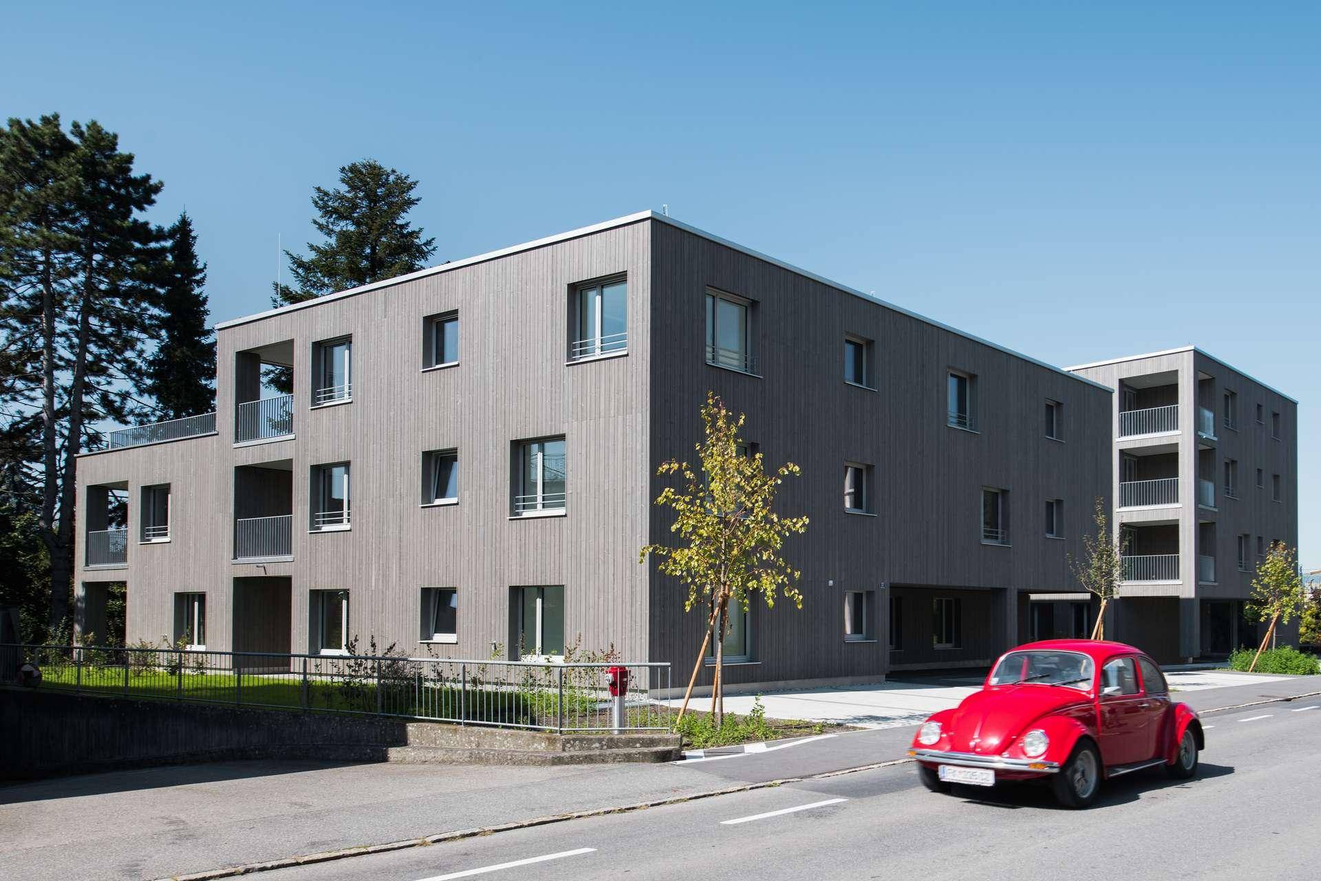 Modernes Mehrfamilienhaus mit Holzfassade und Balkonen, ein roter Käfer fährt vorbei.