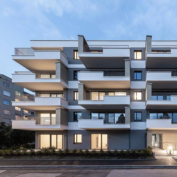 Ansicht eines modernen Mehrfamilienhauses mit gestaffelten Balkonen und beleuchteten Innenräumen am Abend.