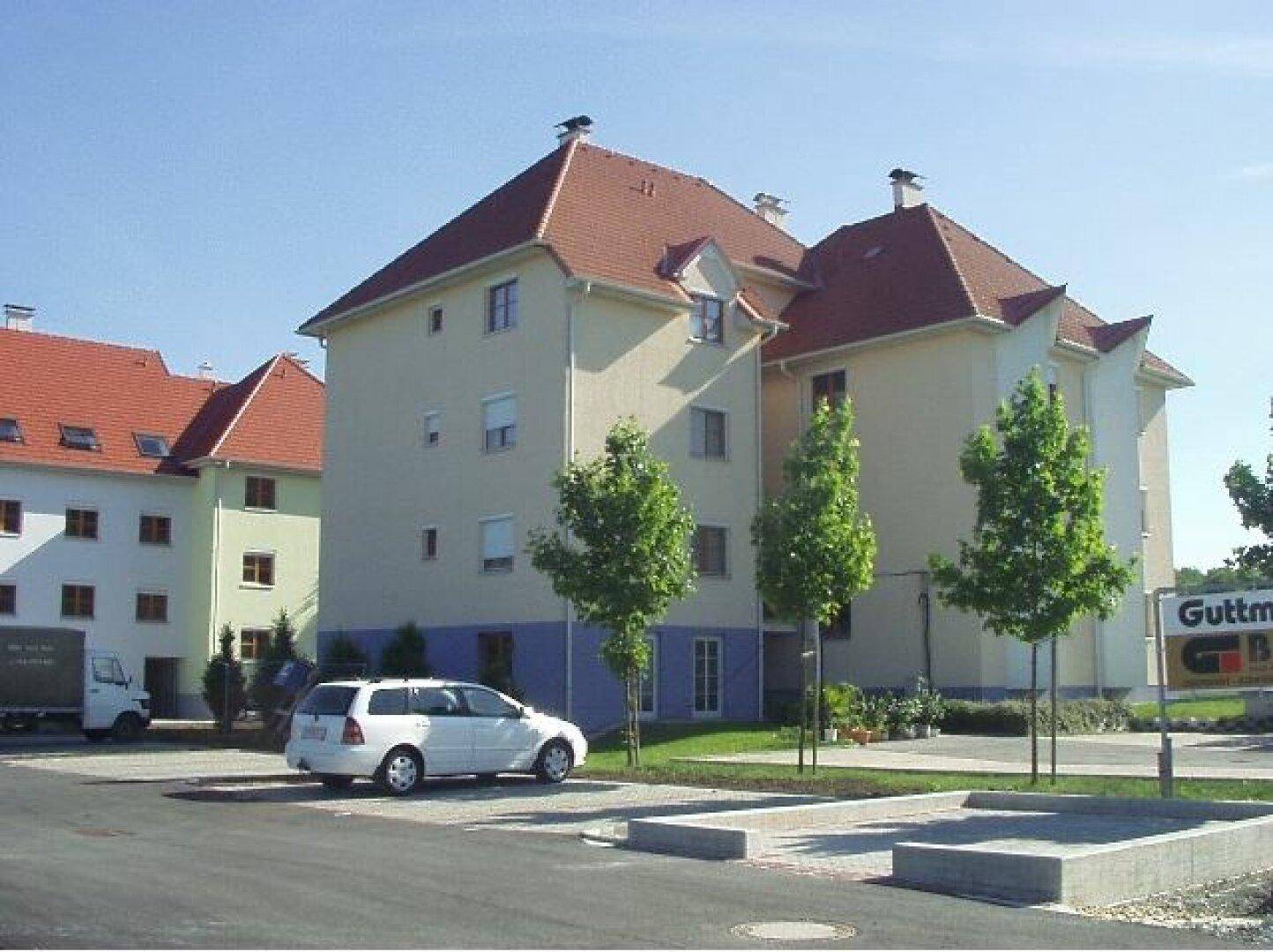 Wohngebäude mit beiger Fassade und rotem Ziegeldach, umgeben von Parkplätzen und jungen Bäumen.