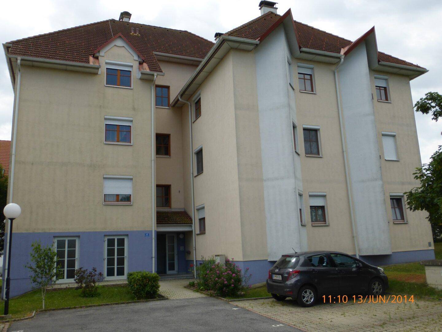 Mehrfamilienhaus mit beiger Fassade, blauer Sockelzone und Satteldach, umgeben von Grünflächen und Parkplätzen.