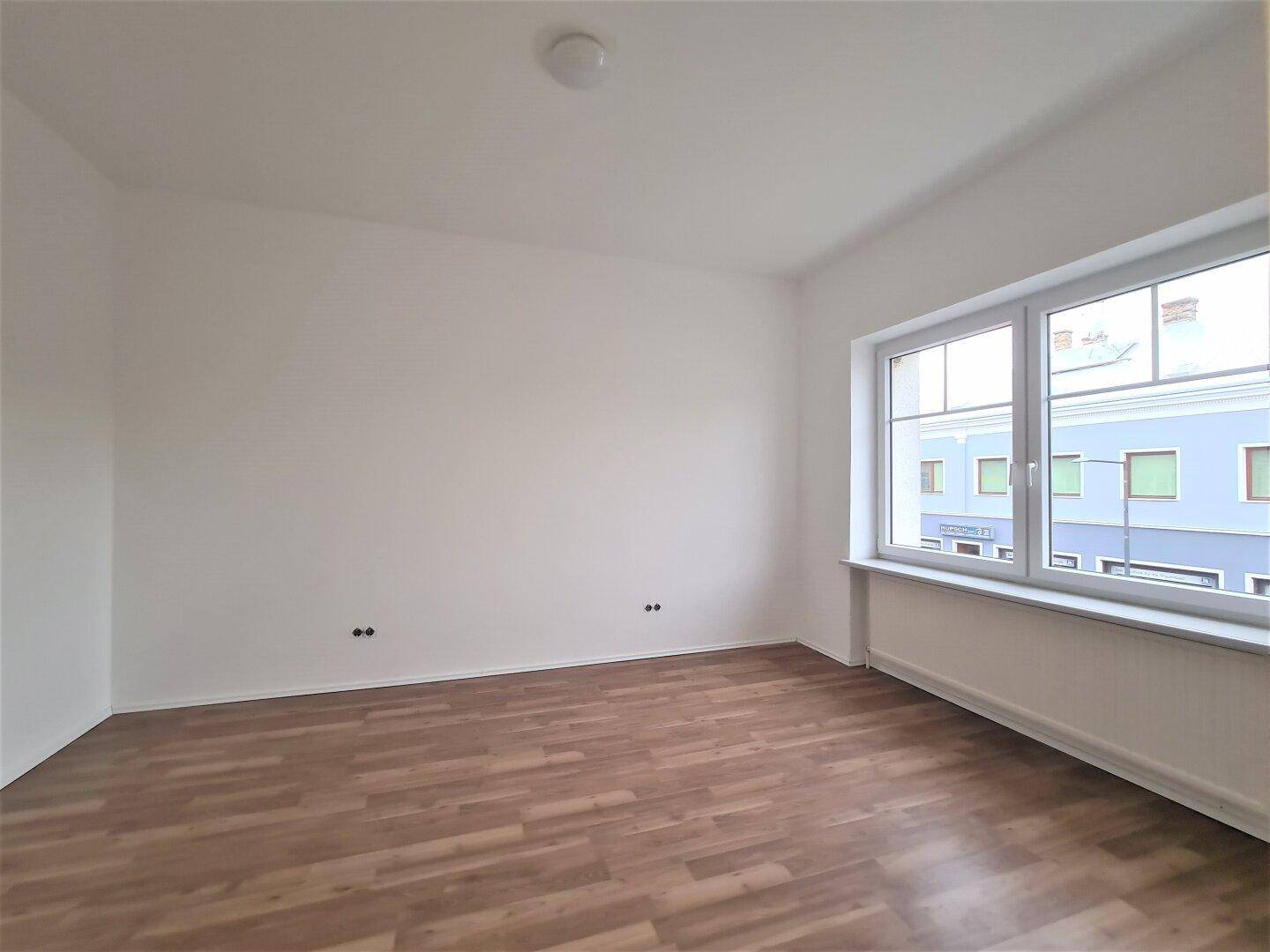 Heller, leerer Raum mit Laminatboden und einem großen Fenster mit Blick auf die Umgebung.