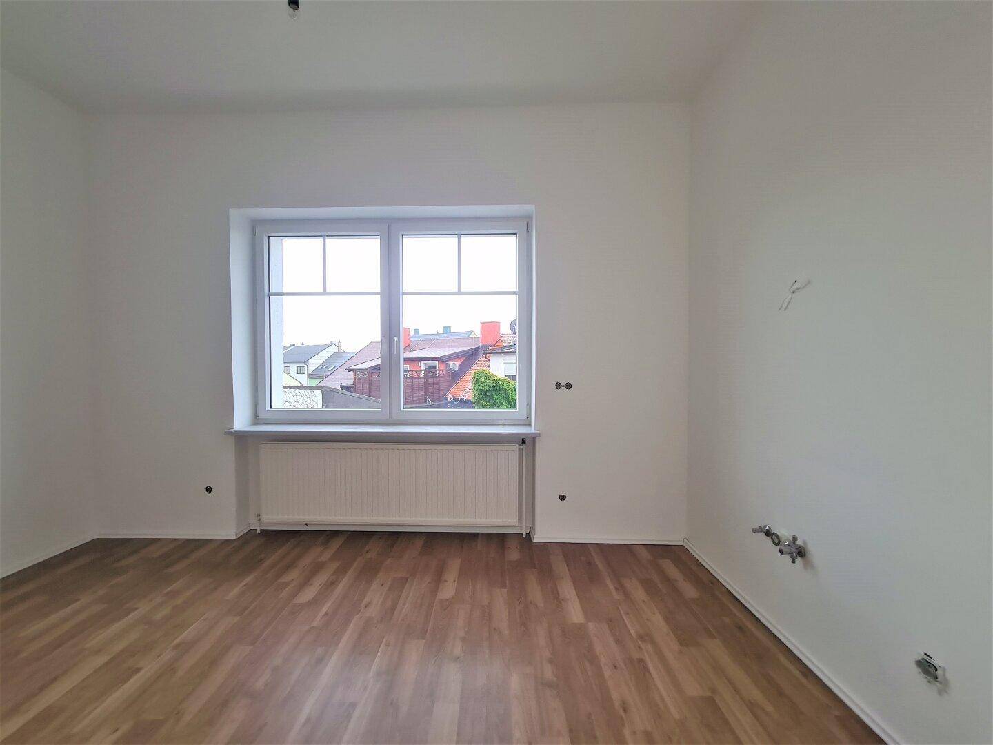 Heller, leerer Raum mit Laminatboden und einem großen Fenster, das viel Tageslicht hereinlässt.