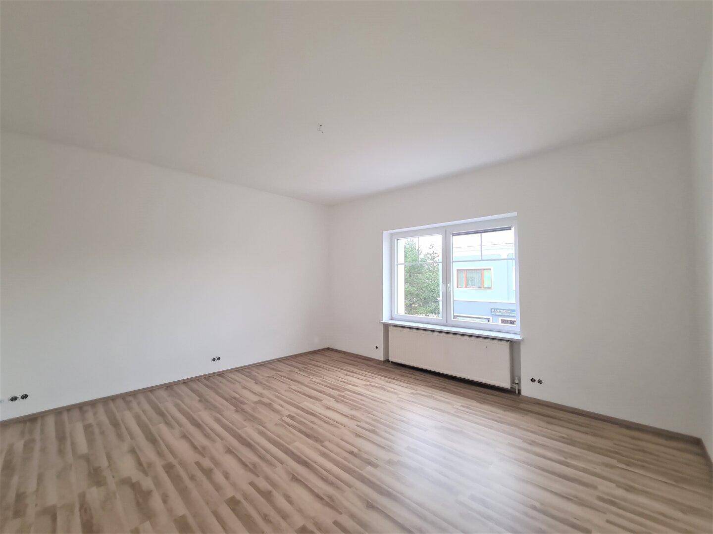 Heller, leerer Raum mit Laminatboden und einem großen Fenster, das eine grüne Aussicht bietet.