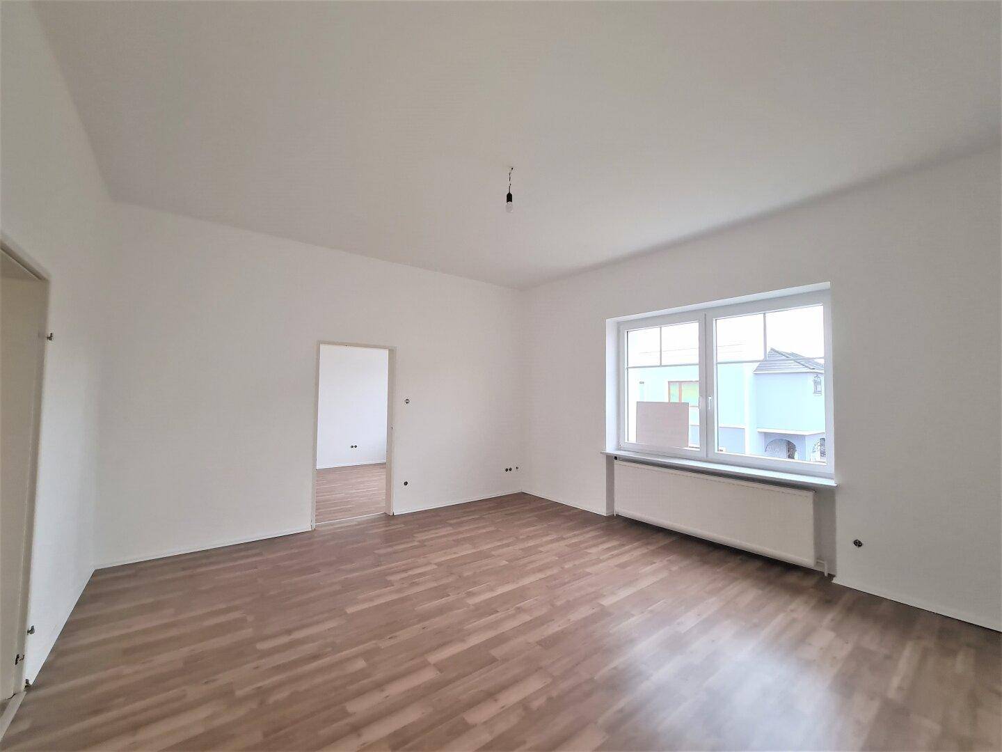 Heller, leerer Raum mit Laminatboden und einem Durchgang zu einem weiteren Zimmer, gut beleuchtet.