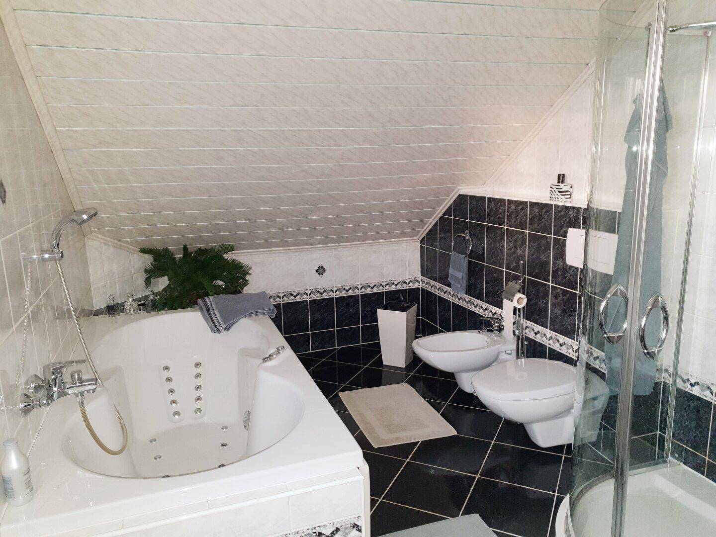 Badezimmer mit Eckbadewanne, Dusche, Bidet und dunklen Fliesen unter Dachschräge.
