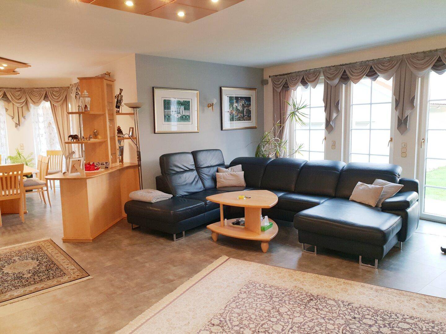 Geräumiger Wohn-Essbereich mit dunklem Ledersofa, Fliesenboden und Zugang zur Terrasse durch große Fenster.
