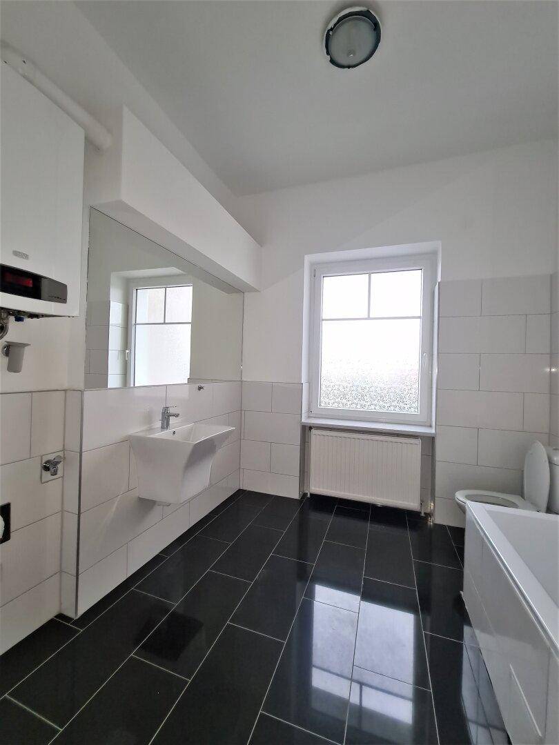 Helles Badezimmer mit Waschbecken, WC, Badewanne und Fenster, ausgestattet mit dunklen Fliesen.