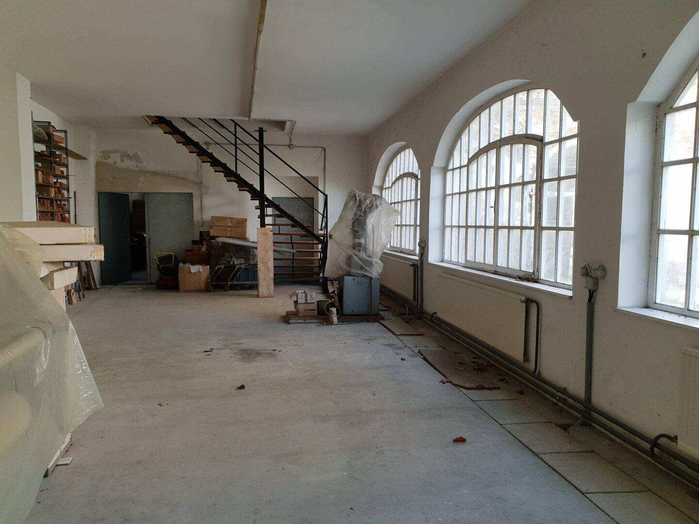Großer, leerer Raum mit Betonboden, hohen Bogenfenstern und einer Metalltreppe zu einer oberen Ebene.