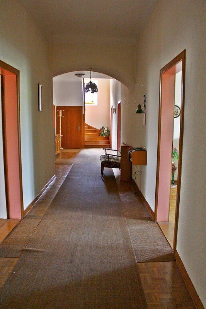 Langer Flur mit Holzparkett, Holztüren und einem eleganten Treppenaufgang im Hintergrund.