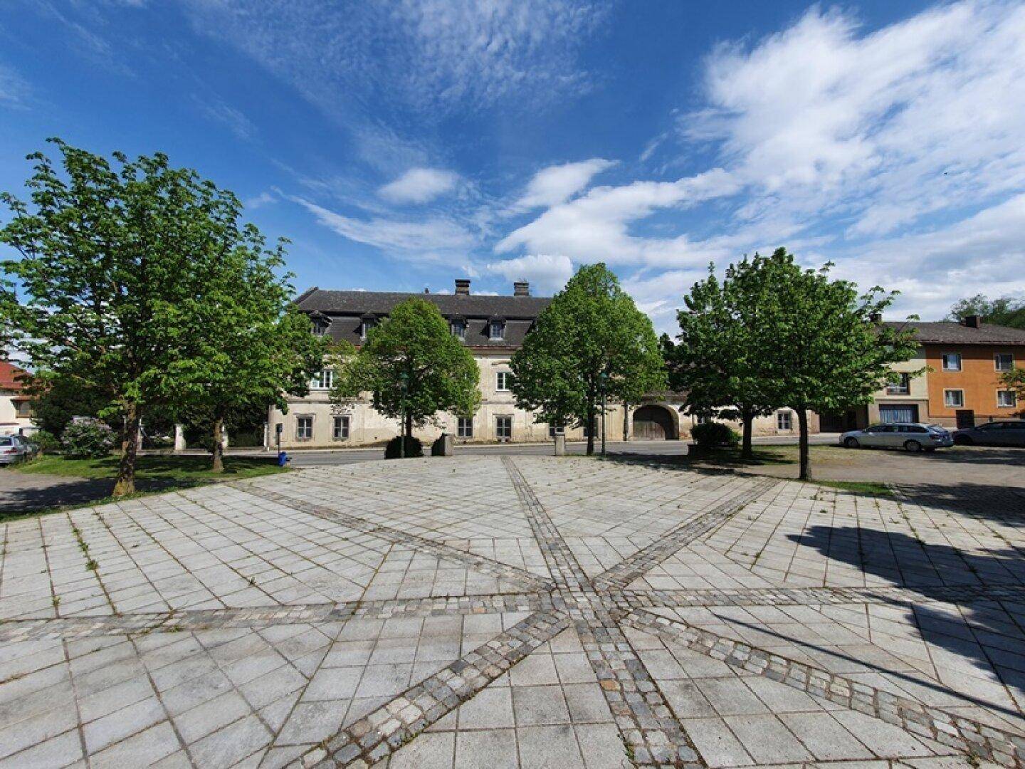 Historisches Gebäude mit gepflastertem Vorplatz und Bäumen unter einem blauen Himmel.