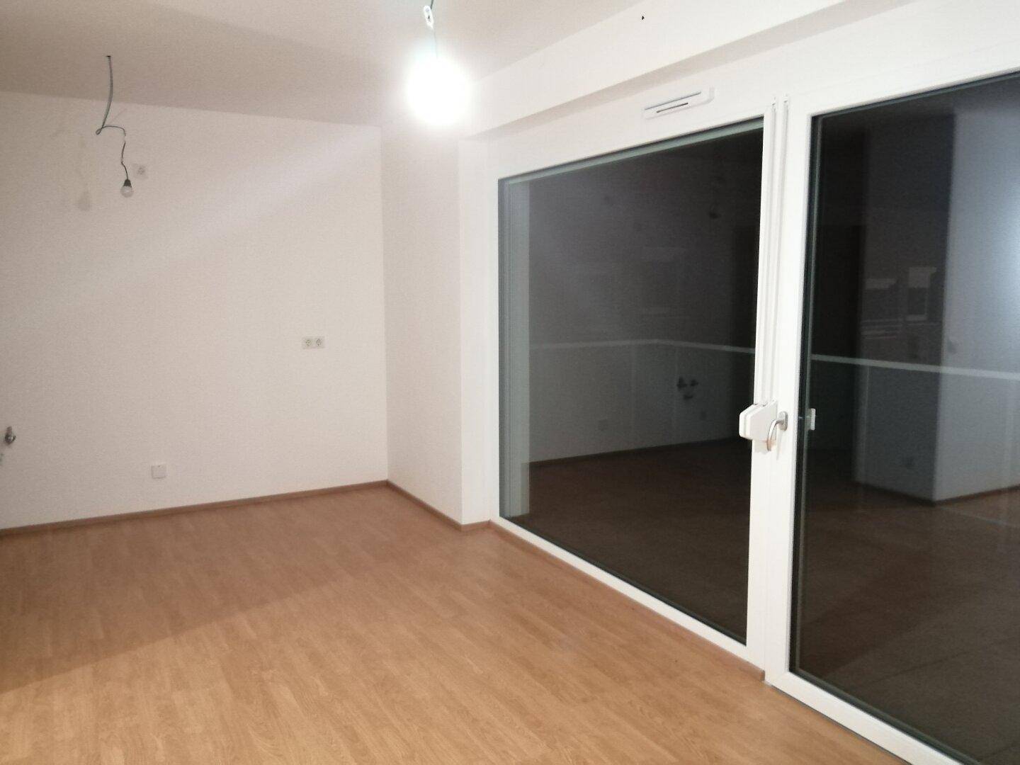 Heller, leerer Raum mit Laminatboden und einer großen Glasschiebetür, die viel Licht hereinlässt.
